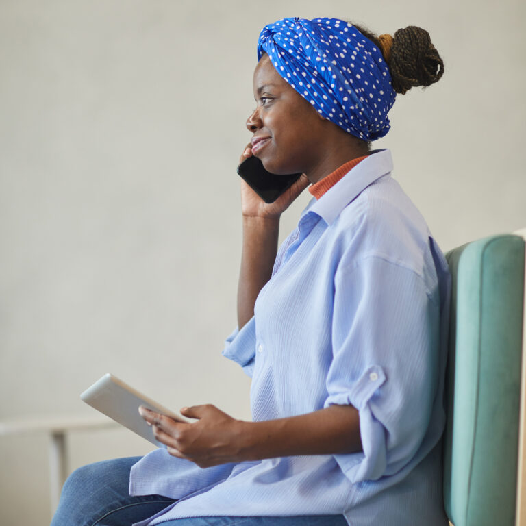 African young businesswoman talking on the mobile phone and using digital tablet while sitting on sofa